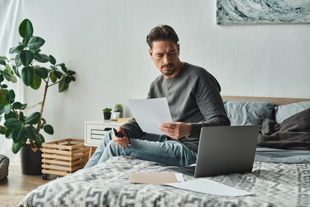 focused and bearded man holding smartphone and using laptop near documents on bed, remote work - Photo, Image