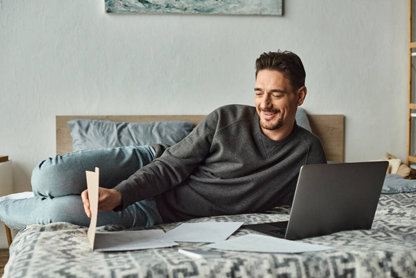 веселый и бородатый мужчина с помощью ноутбука во время работы удаленно из дома в спальне, документы - Фото, изображение