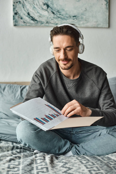 hombre barbudo en auriculares inalámbricos analizando gráficos mientras escucha música en el dormitorio, el trabajo desde casa - Foto, Imagen