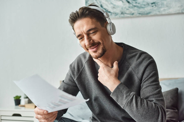 glücklicher Mann mit drahtlosen Kopfhörern, der Grafiken analysiert und im Schlafzimmer Musik hört, von zu Hause aus arbeitet - Foto, Bild