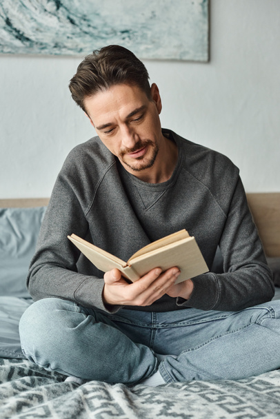 γενειοφόρος άνδρας σε γκρι casual jumper ανάγνωση βιβλίο, ενώ χαλαρώνοντας το Σαββατοκύριακο στην κρεβατοκάμαρα, αναψυχής - Φωτογραφία, εικόνα