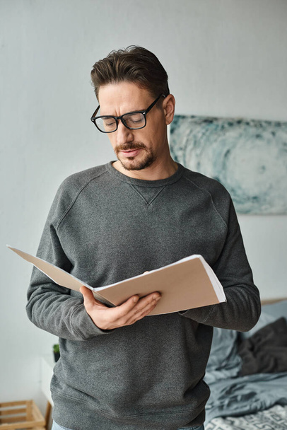 soustředěný muž v brýlích a svetru analyzující dokumenty při práci z domova v ložnici - Fotografie, Obrázek