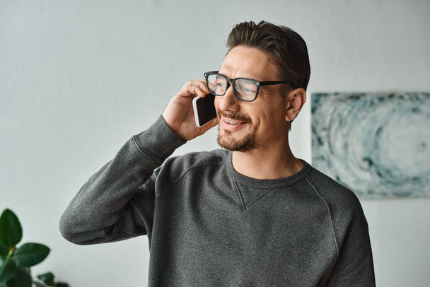 bearded man in eyeglasses and grey jumper talking on smartphone in modern bedroom, phone call - Fotó, kép