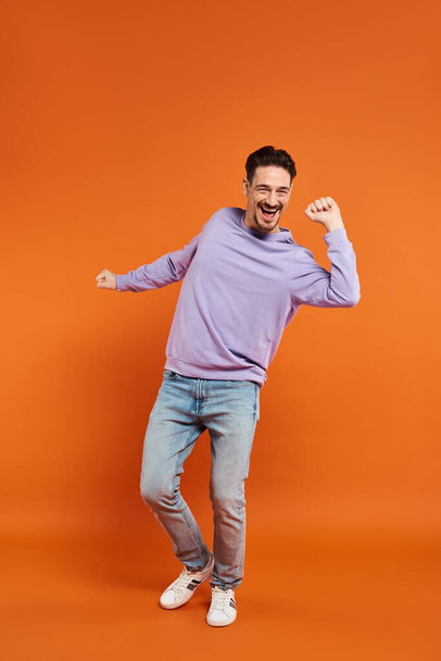 longitud completa de hombre excitado y barbudo en suéter púrpura y pantalones vaqueros bailando sobre fondo naranja - Foto, imagen