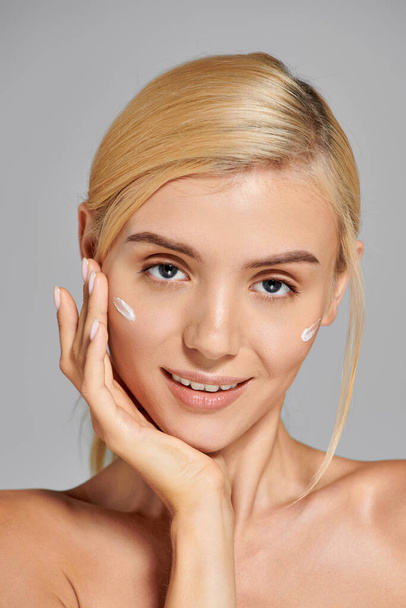 attrayant jeune femme avec confiant regardant l'application de crème sur ses joues sur fond gris - Photo, image