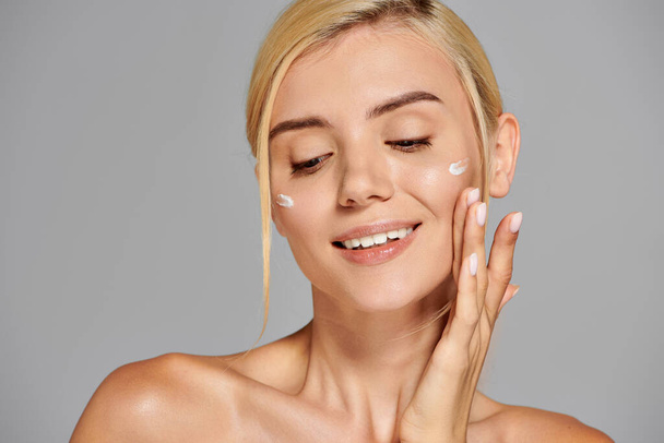 заманчивая женщина в возрасте 20 лет со здоровой кожей счастлива наносить крем на лицо на сером фоне - Фото, изображение