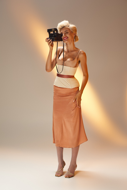 glamourous γυναίκα 20 ετών σε κομψό ντύσιμο λήψη φωτογραφιών με ρετρό κάμερα σε γκρι φόντο - Φωτογραφία, εικόνα
