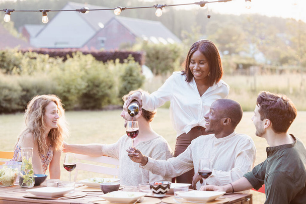 Une femme noire se tient debout en train de verser du vin avec un sourire, tandis que ses amis, une femme caucasienne et deux hommes, l'un noir et l'autre caucasien, regardent avec des lunettes à la main. Ils sont assis autour d'une table d'extérieur rustique - Photo, image