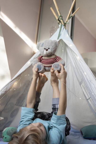 Çocuk, çocuk odasında bir çadırda uzanmış oyuncak ayıyla oynarken mutlu bir şekilde ayaklarını kaldırıyor. fantezi, ilginç çocukluk, yumuşak oyuncaklarla oynamak, neşe - Fotoğraf, Görsel
