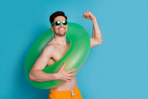 Photo portrait of nice young male raise fist float ring muscle wear trendy yellow shorts isolated on blue color background summer vacation. - 写真・画像