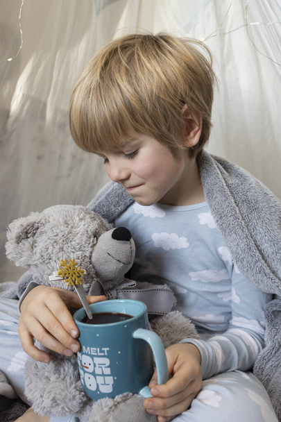 petit garçon caucasien en pyjama assis dans une wigwam dans une chambre d'enfants, recouvert d'une couverture, étreint un ours en peluche et tient une tasse de chocolat chaud. Enfance, jouer avec les jouets, tendresse - Photo, image