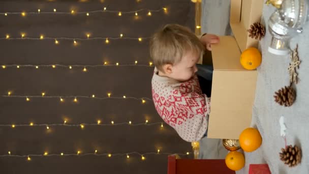 Mały zabawny chłopiec chowa się w kartonowym pudełku i wydziera z niego, uśmiechając się. Boże Narodzenie dzieci - Materiał filmowy, wideo