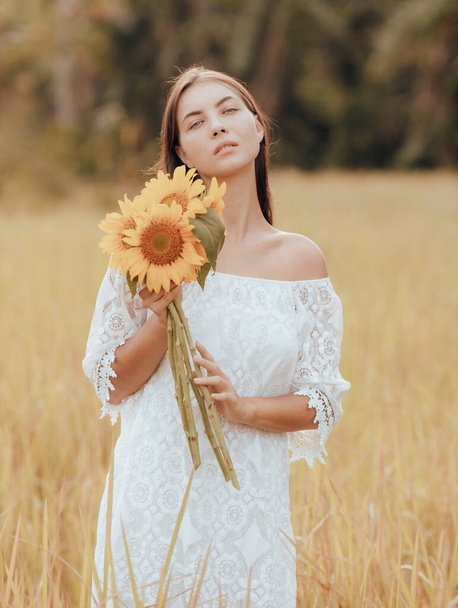 Junge Frau, die auf einem Feld spaziert und einen Strauß Sonnenblumen in der Hand hält. Porträt einer kaukasischen Frau im weißen Kleid. Sommerferien. Vertikale Anordnung. Lifestyle-Konzept. Romantische Stimmung. - Foto, Bild