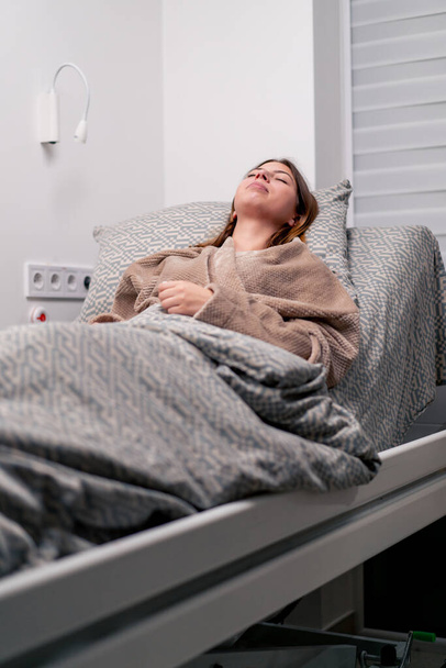 ένας άρρωστος ασθενής σε ένα νοσοκομείο είναι ξαπλωμένος σε ένα κρεβάτι φτερνίζεται και υποφέρει από συνάχι βήχα και πονοκεφάλους - Φωτογραφία, εικόνα