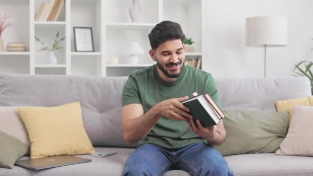 Boldog indián férfi alkalmi öltözékben ül szürke kanapén könyvvel a kezében, és jól látható jelet mutat. Vonzó fiatal férfi elégedett az új irodalmat olvasni otthon. - Felvétel, videó