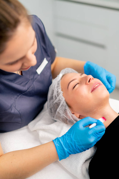 врач-косметолог в перчатках держит кисть и наносит маску здоровья на лицо и шею клиентки в салоне красоты - Фото, изображение