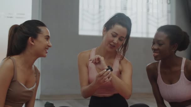 Medium shot di gruppo multiculturale di atlete che discutono la nuova app fitness su smartwatch allo stretching studio - Filmati, video