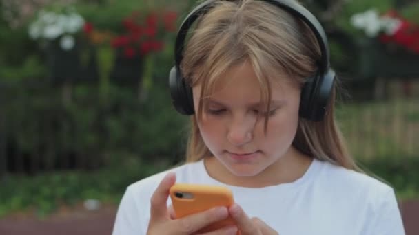 街の背景とスマートフォンを使用して立っている黒いスタイリッシュなヘッドフォンを持つ美しい若い十代の少女. 女の子が携帯電話でロケーションアプリを閲覧しています. 高品質の4k映像 - 映像、動画