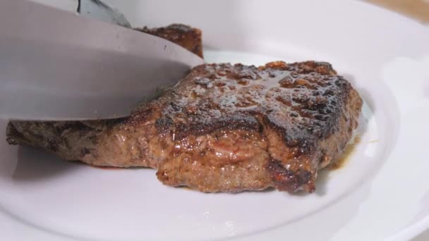 Un grosso pezzo di bistecca di manzo appena grigliata su un piatto. Carne arrosto media con sangue al rallentatore. Un coltello e una forchetta tagliano un pezzo vista da vicino - Filmati, video