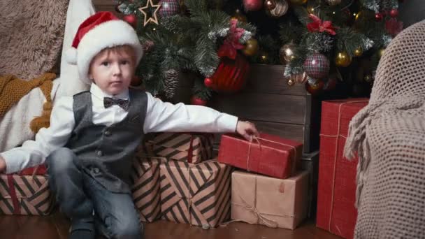 Glücklicher kleiner Junge mit Nikolausmütze prüft Geschenke unter dem Tannenbaum. Kind öffnet Geschenkbox an Heiligabend. Weihnachtskonzept - Filmmaterial, Video