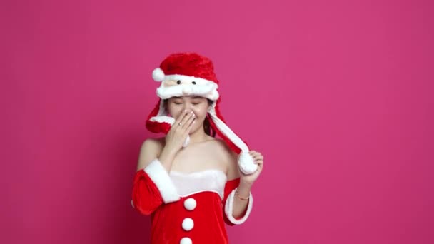 Filmación de una mujer asiática vestida con tela temática navideña jugando con su sombrero tímidamente y un fondo rosa en la espalda - Imágenes, Vídeo