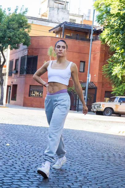 молодая модная женщина переходит улицу в короткой белой футболке и серых штанах для пробежки. у нее есть фитнес-тело. - Фото, изображение