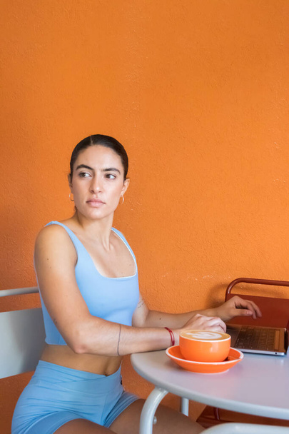 beautiful young woman enjoys a cup of coffee while working on her computer. she is wearing gray clothes and has a serious look on her face. - Photo, Image