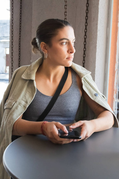 attraktive Frau, die mit ihrem Handy in einem Café sitzt. braunhaarige Frau in beiger Jacke - Foto, Bild