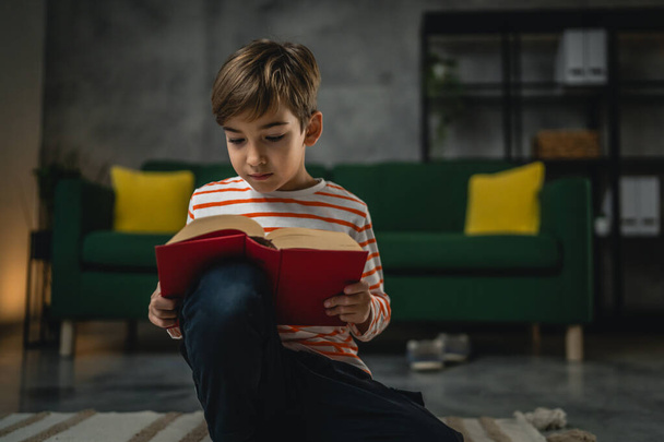 Ένα καυκάσιο αγόρι στο σπίτι διαβάζει ένα βιβλίο στο μπροστινό μέρος της βραδιάς αντιγραφή χώρο πραγματικό άνθρωποι εκπαίδευση έννοια - Φωτογραφία, εικόνα