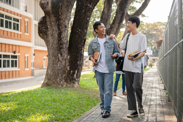 Dwóch wesołych azjatyckich studentów cieszy rozmowa podczas wspólnego chodzenia do szkoły, chodzenie po ścieżce na kampusie. Koncepcja przyjaźni - Zdjęcie, obraz