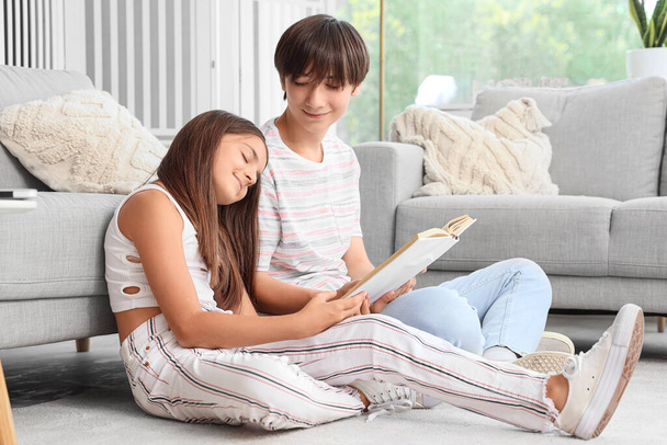 Linda chica con su hermano sentado en el suelo y leyendo libro en casa - Foto, Imagen