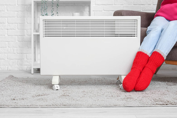 Νεαρή γυναίκα με ζεστές χειμερινές κάλτσες κοντά σε ηλεκτρική θερμάστρα. Έννοια της εποχής θέρμανσης - Φωτογραφία, εικόνα