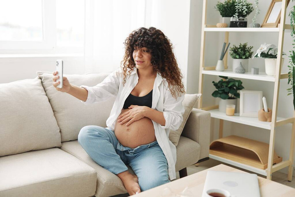 La bloguera embarazada se sienta en el sofá de su casa y se toma fotos por teléfono, selfies y videollamadas, consulta con el médico en línea, manejo del embarazo. Foto de alta calidad - Foto, imagen