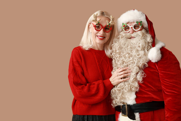 Happy Santa Claus and beautiful mature woman in Christmas glasses on brown background - Photo, Image