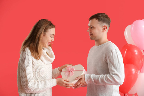 Preciosa pareja con caja de regalo y globos en forma de corazón sobre fondo rojo. Celebración de San Valentín - Foto, imagen