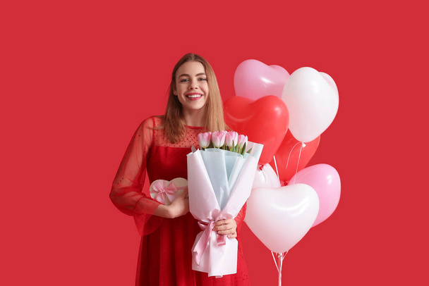 Jolie jeune femme avec coffret cadeau, bouquet de fleurs et ballons en forme de coeur sur fond rouge. Fête de la Saint-Valentin - Photo, image