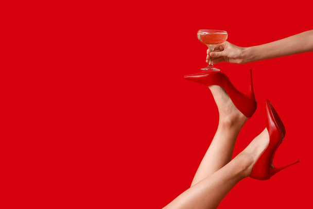 Женские ноги в туфлях на высоком каблуке с бокалом шампанского на красном фоне. Новогодняя вечеринка - Фото, изображение