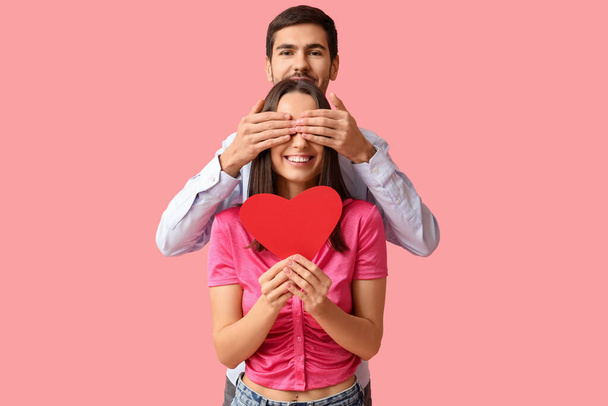 Красивая молодая пара с красным бумажным сердцем на розовом фоне. Празднование Дня Святого Валентина - Фото, изображение