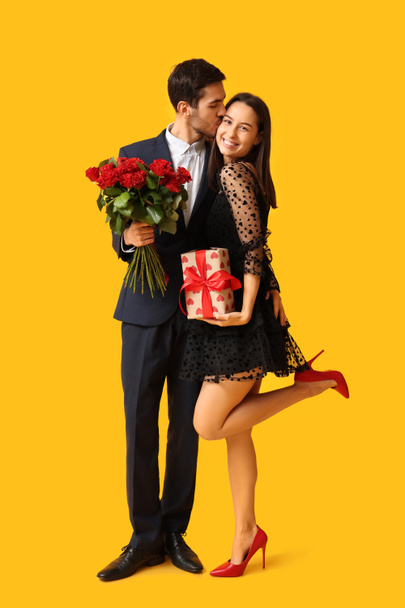 Junger Mann küsst seine Freundin mit einem Strauß roter Rosen und einer Geschenkbox auf gelbem Hintergrund. Valentinstag-Feier - Foto, Bild