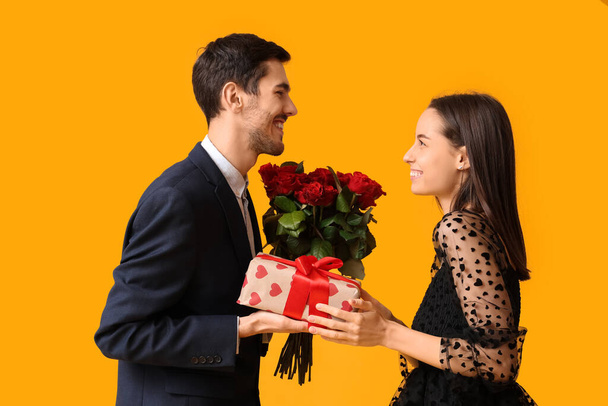Όμορφο νεαρό ζευγάρι με μπουκέτο κόκκινα τριαντάφυλλα και κουτί δώρου σε κίτρινο φόντο. Γιορτή του Αγίου Βαλεντίνου - Φωτογραφία, εικόνα