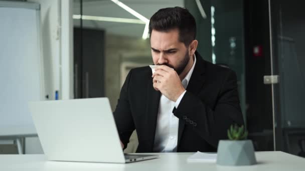 Displeased handsome male coughing and blowing nose in paper tissue while sitting at table with modern laptop. Ill caucasian businessman having running nose and sore throat at bright modern office. - Footage, Video
