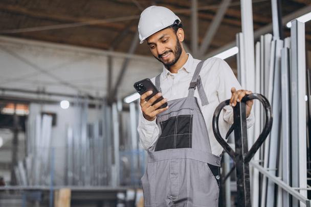 Portrait of a happy Hindu worker in a white helmet and overalls holding a hydraulic truck and talking on the phone against a background of a factory and aluminum frames. - Photo, Image