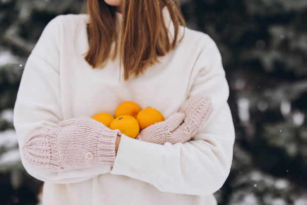 Une belle fille heureuse en tricot blanc tient des mandarines en hiver sur un fond de pin - Photo, image