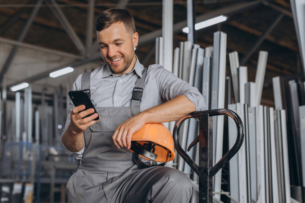 Retrato de un trabajador feliz en un casco naranja y overoles sosteniendo un camión hidráulico y hablando por teléfono sobre un fondo de una fábrica y marcos de aluminio. - Foto, Imagen