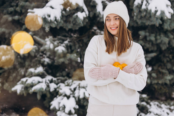 Красивая счастливая девушка в белой трикотажной одежде держит мандарины зимой на фоне сосны - Фото, изображение