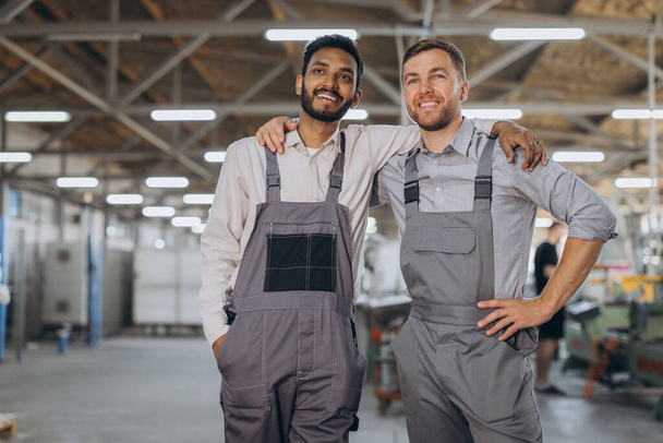 Kaksi onnellista kansainvälistä työntekijää haalareissa halaavat toisiaan tehdastaustaa vasten - Valokuva, kuva