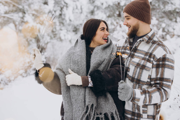 Glimlachend romantisch paar in gebreide hoeden poseren met sterretjes in het winterbos, Bengaalse lichten in handen houden, kerstvakantie samen vieren, ruimte kopiëren - Foto, afbeelding