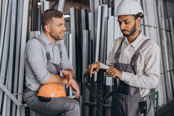 Portret dwóch międzynarodowych pracowników w twardych kapeluszach wykonujących przerwę w pracy i odpoczywających rozmawiających ze sobą na tle fabrycznym z przestrzenią do kopiowania - Zdjęcie, obraz