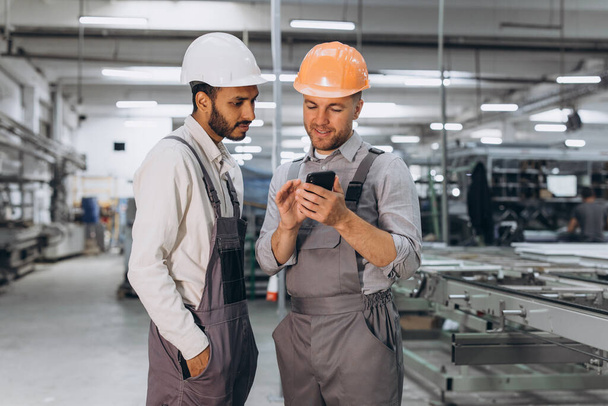 Two international male workers in overalls looking at the phone against the background of the production of aluminum PVC windows and doors - Photo, Image