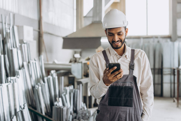 Ευτυχισμένος γενειοφόρος Ινδός εργάτης σε εργοστάσιο με ειδική στολή και λευκό σκληρό καπέλο κρατώντας το τηλέφωνο και έχοντας διάλογο στην παραγωγή μεταλλικών-πλαστικών παραθύρων και θυρών - Φωτογραφία, εικόνα